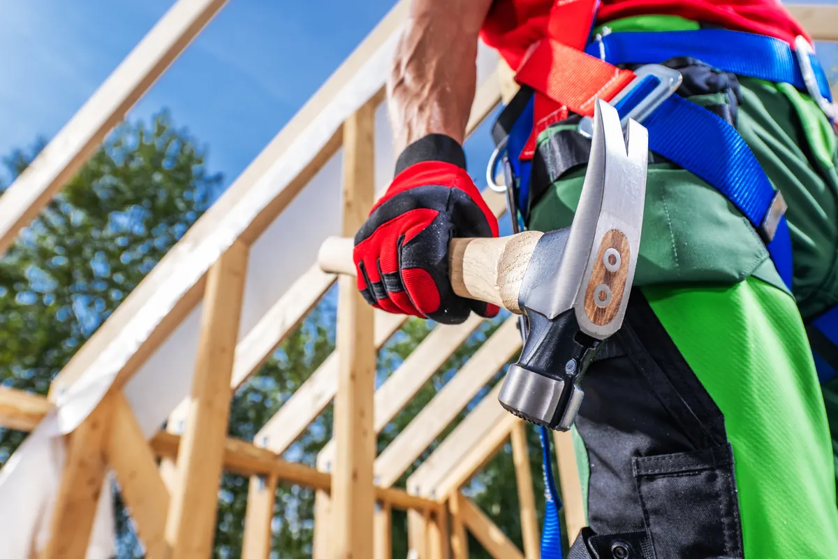 Zimmermann mit Hammer am Gürtel vor einem neuen Dachstuhl aus Holz